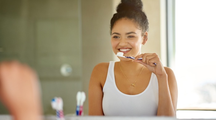 Young woman with her hair in a bun wearing a white tank top and brushing her teeth in front of a mirror.