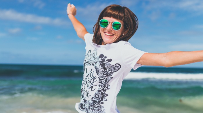 Brunette woman wearing a white t-shirt and sunglasses jumps into the air, arms outstretched, by the ocean