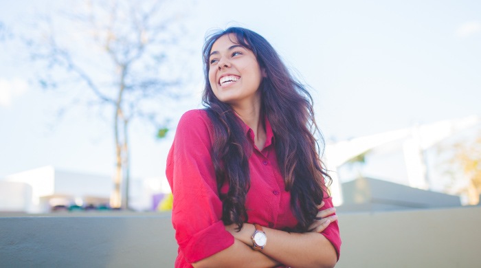 Brunette woman wearing a red blouse and a watch folds her arms and smiles as she looks to the side in a parking lot