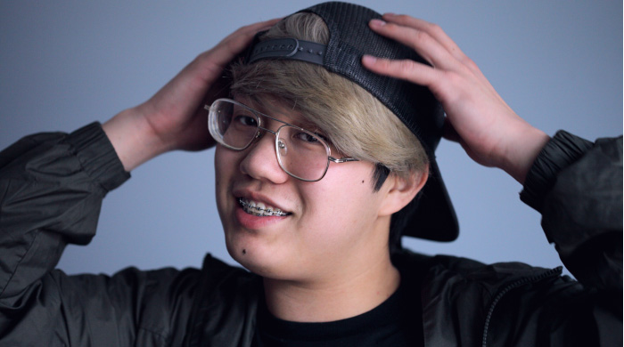 boy with blonde hair, glasses, braces on his teeth, wearing a backwards baseball cap, with his hands on his head.