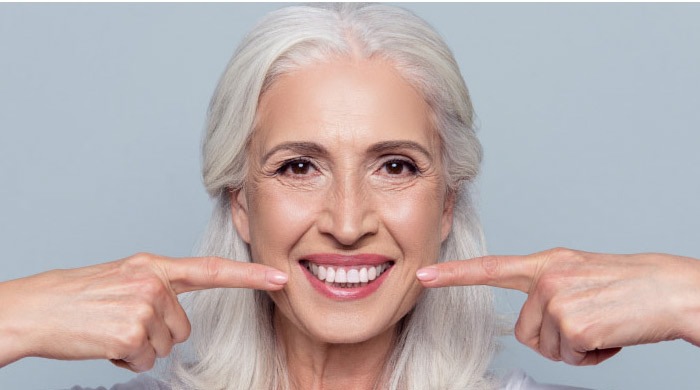 woman with long white hair smiles and points to her white teeth with both forefingers