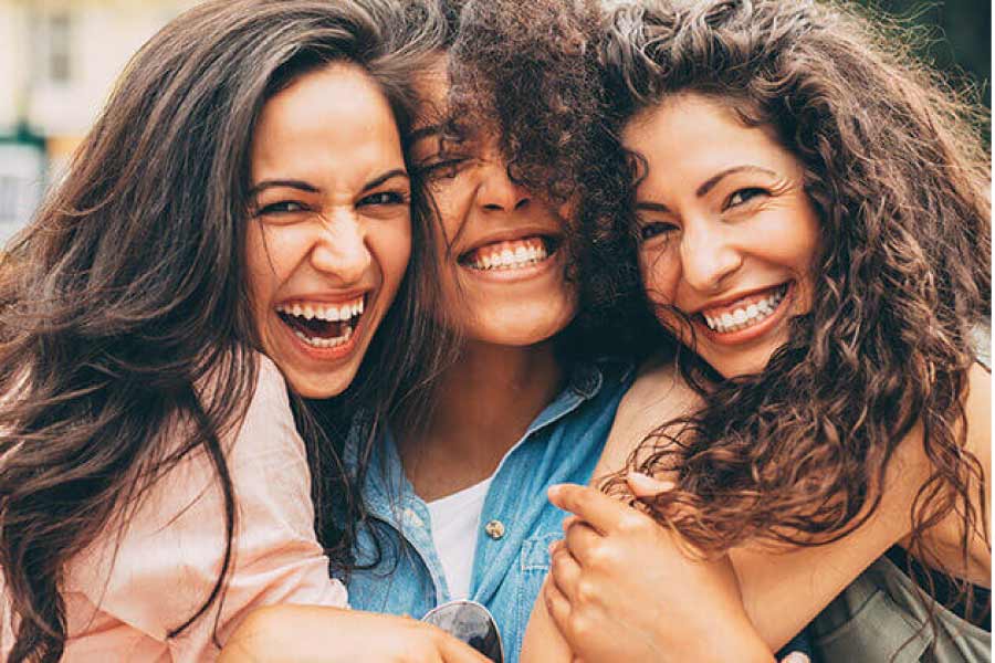 three brown haired young women hug and smile showing off their teeth